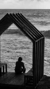 A person sits at the end of a dock, back to the camera. They are wearing a hoodie and appear to be contemplating past traumas as they watch the ocean waves.